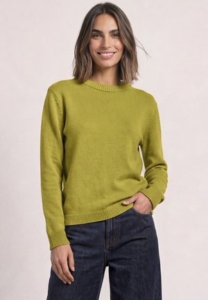 Femme aux cheveux bruns mi-longs portant un pull vert citron et un jean bleu foncé, souriante, une main dans la poche sur un fond uni.