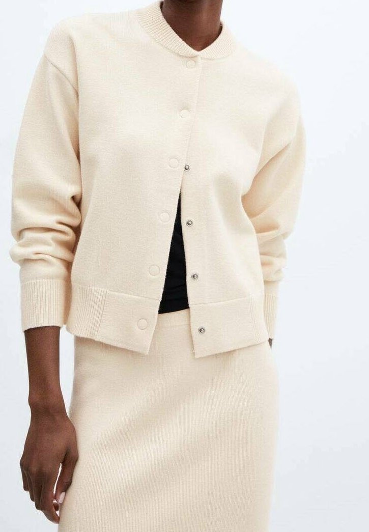 Personne portant un cardigan beige boutonné et une jupe assortie avec un haut noir en dessous sur un fond blanc uni.