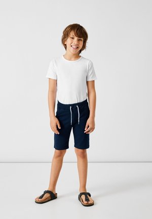 Weißes T-Shirt mit kurzen Ärmeln und marineblauen Joggingshorts mit Kordelzug, dazu schwarze Flip-Flops, stehend vor einem neutralen Hintergrund.