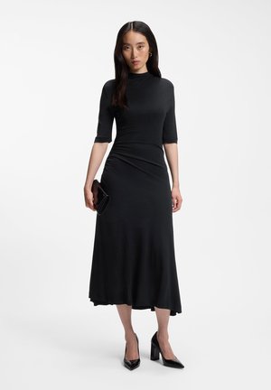 Femme portant une robe midi noire avec des manches jusqu'au coude, tenant une pochette noire et portant des chaussures à talons noirs sur fond blanc.