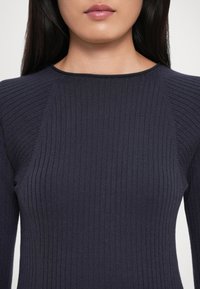 Žena v tmavě námořnické ribbed dlouhé mikině s kulatým výstřihem, ukazující rty a dolní část obličeje zblízka.