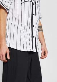 Camicetta bianca a maniche corte con strisce verticali nere, apertura con bottoni e rifiniture nere a contrasto lungo la pattina. Pantaloni neri visibili sotto.