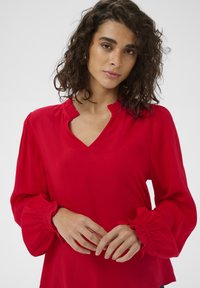 Blouse rouge à manches longues avec un col en V et un col volanté. Caractéristiques : poignets élastiqués et texture de tissu lisse.