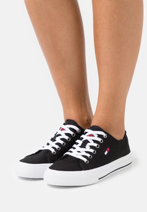 Sorte lave canvas-sneakers med hvide snørebånd og såler, båret på fødder mod en ensfarvet hvid baggrund.