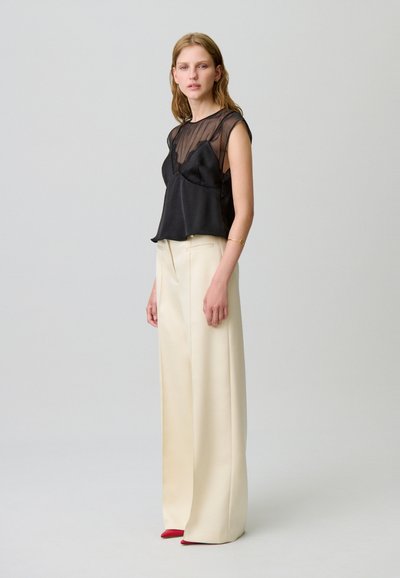 Mujer de pie con pantalones anchos crema, top sin mangas negro de gasa y satén, y zapatos de punta rojos, frente a un fondo liso de color gris claro.