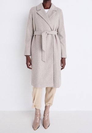 Vrouw draagt een beige wollen jas met riem over lichte broek en puntige beige enkellaarzen, staand tegen een effen witte achtergrond.