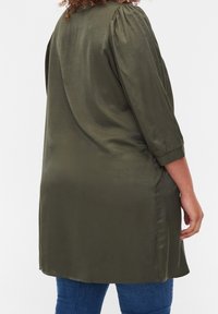 Tunique vert olive à manches longues avec détail froncé aux épaules et fentes latérales. Tissu lisse et léger. Portée sur un jean bleu.