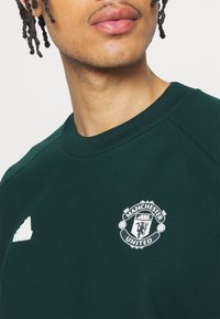 Grön sweatshirt i mjukt tyg med en vit Manchester United-logotyp och en vit Adidas-emblem på vänster bröst.