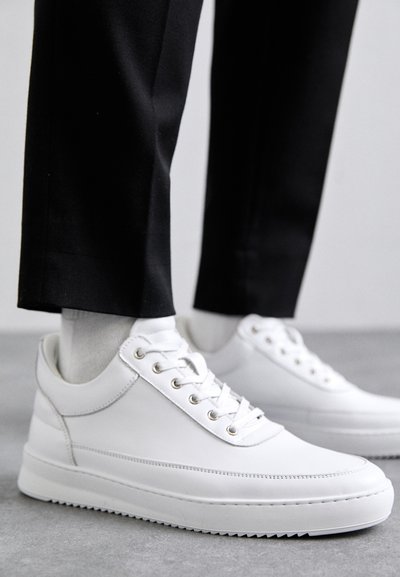 Weiße Leder-Sneaker mit flacher Gummisohle, ausgestattet mit einer glatten Oberfläche, runder Zehenpartie und Metallösen für die Schnürsenkel. Getragen mit schwarzen Hosen.