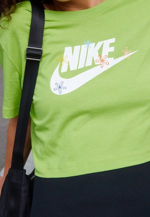 Osoba nosząca limonkowo-zieloną krótka koszulkę Nike z kolorowymi kwiatowymi wzorami oraz widocznym czarnym paskiem torby na ramię.