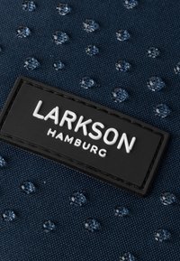 Tengerészkék anyag vízcseppekkel és texturált felülettel. Egy fekete, téglalap alakú címke található rajta fehér dombornyomott szöveggel: "LARKSON HAMBURG."