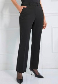 Vrouw in zwarte op maat gemaakte broek en zwarte hoge hakken, staande met één hand in de zak tegen een witte paneelwand.