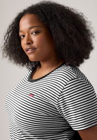 Ung kvinne med naturlig krøllete hår iført en svart-hvit stripete Levi's T-skjorte, som ser selvsikkert over skulderen.