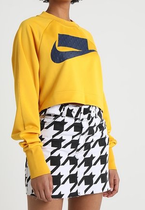 Personne portant un sweat court jaune avec un logo Nike noir et une jupe taille haute pied-de-poule noir et blanc.