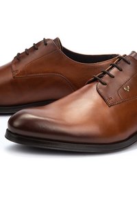Une paire de chaussures de ville en cuir brun poli avec des lacets noirs et des semelles noires basses sur un fond blanc.