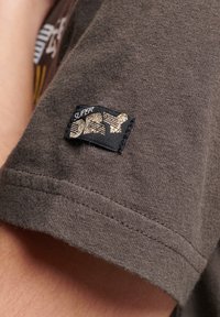 Brązowy bawełniany t-shirt z rękawem, na którym znajduje się czarna metka ze złotym napisem oraz grafiką. Tkanina ma miękką, teksturowaną powierzchnię.