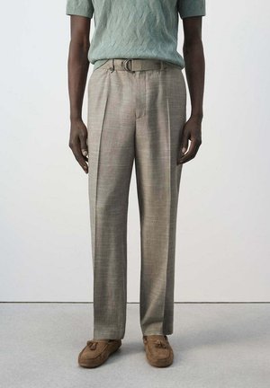 Uomo con pantaloni beige plissettati, mocassini marroni con nappine e maglione verde a maniche corte in piedi davanti a uno sfondo uniforme.