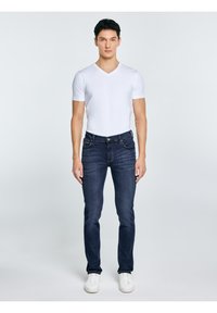 Bărbat în picioare, orientat frontal, purtând un tricou alb cu guler în V, blugi întunecați și adidași albi, pe un fundal simplu și deschis la culoare.