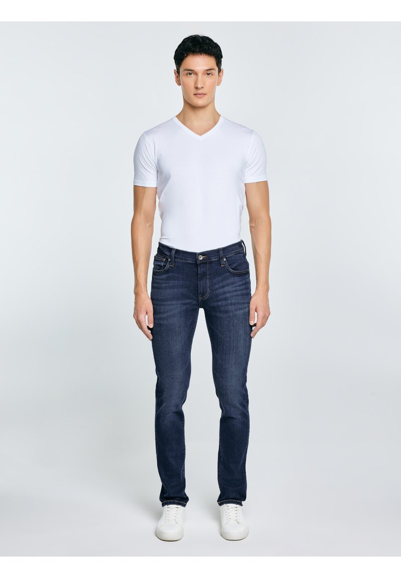 Bărbat în picioare, orientat frontal, purtând un tricou alb cu guler în V, blugi întunecați și adidași albi, pe un fundal simplu și deschis la culoare.