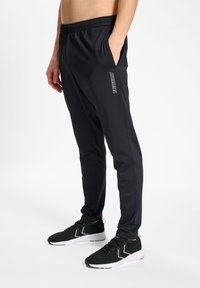 Black athletic pants with a fitted design, elastic waistband, side pockets, and a reflective logo detail on the left leg. Worn with black sneakers.