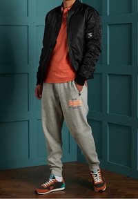 Personne portant une veste bomber noire, un polo orange, un pantalon de survêtement gris avec un logo, et des chaussures de sport multicolores, se tenant contre un mur en panneaux teal.