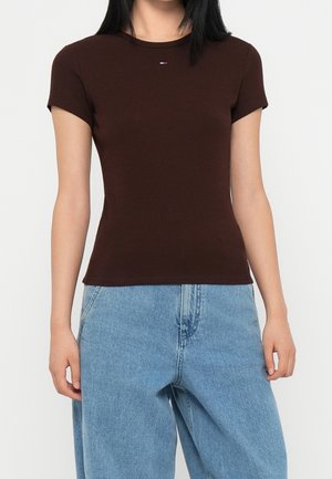 Kobieta nosząca dopasowaną brązową bluzkę z krótkim rękawem i małym logo na piersi oraz luźne jasnoniebieskie jeansy.