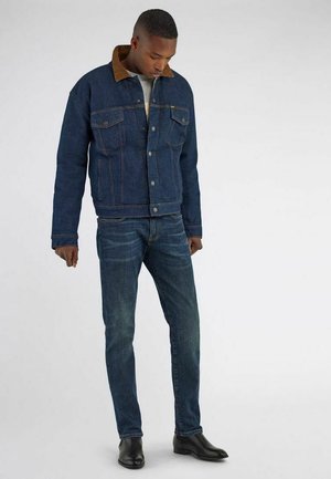 Uomo che indossa una giacca di jeans blu scuro con colletto marrone, jeans abbinati, stivali neri, in piedi davanti a uno sfondo grigio uniforme.