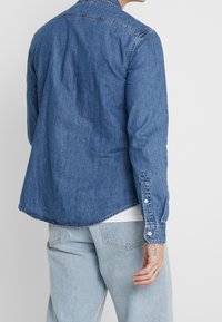 Tummansininen denim-paita, pitkähihaisena, kauluksella ja napillisilla hihansuilla. Ominaisuuksiin kuuluu kaareva helma ja sileä pinta. Paita on päällysvaatteena vaaleiden farkkujen päällä.