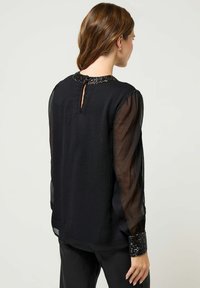 Blusa nera con maniche lunghe trasparenti, tessuto testurizzato e colletto con perline e apertura a forma di chiave sul retro. Maniche con polsini e dettagli in paillettes.