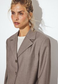 Blazer ajusté marron clair avec un col cranté, deux boutons et des épaules structurées, fabriqué en tissu texturé pour un look professionnel.