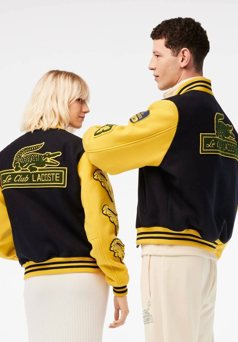 Veste varsity Lacoste avec un corps en laine noire, des manches jaunes, des logos crocodile brodés, et des poignets et un ourlet côtelés rayés.