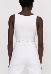 Top blanco sin mangas con un escote redondeado, combinado con jeans blancos de cintura alta que tienen una etiqueta en la parte posterior, costuras visibles y bolsillos.