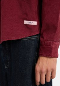 Kordhemd in tiefburgunder mit vertikalen Rippungen, ausgestattet mit einem Etikett, das "ELEMENT CO. SINCE 1992" am Saum trägt. Getragen mit dunkler Jeans.