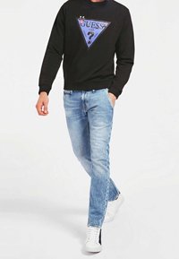 Sudadera negra con un gráfico triangular azul que presenta la palabra "GUESS" y un diseño divertido, combinada con jeans de mezclilla azul claro y zapatillas blancas.