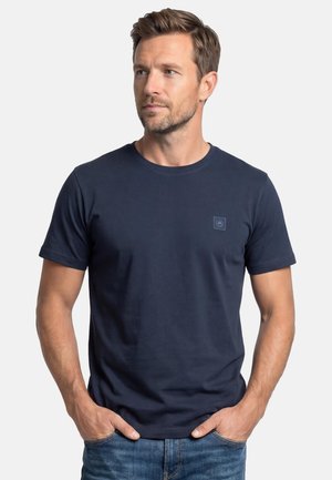 Mann mit kurzen braunen Haaren und leichtem Bart, trägt ein marineblaues kurzärmeliges T-Shirt und blaue Jeans, Hände in den Taschen, blickt nach links.