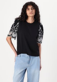 Svart t-shirt med kort ärm och intrikata vita broderade mönster på ärmarna, med en avslappnad passform och mjukt tyg. Bärs tillsammans med ljusblåjeans.