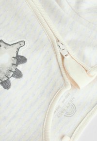 Body en coton avec des rayures jaunes claires et blanches, orné d'une application de tortue grise et d'une fermeture éclair blanche sur le côté. Texture douce.