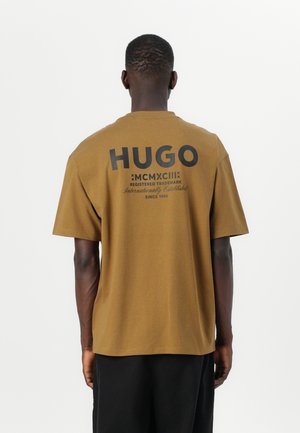 Man shown from behind wearing a brown T-shirt with black "HUGO" and text printed on the upper back against a plain white background.