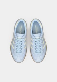 Ανοιχτό μπλε σουέτ αθλητικό παπούτσι με τρεις κάθετες λευκές ρίγες, επίπεδα κορδόνια και υφασμένη καφέ ρητίνη στη σόλα. Κλασικό λογότυπο Adidas στη γλώσσα.