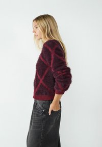 Burgundowy, puszysty sweter o diamentowym wzorze w miękkiej strukturze, z prążkowanymi mankietami i dołem, w połączeniu z czarnymi dżinsami.