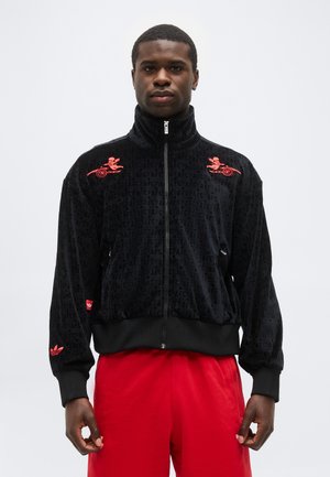 Joven con chaqueta negra con cremallera y logotipos rojos en los hombros y la manga, combinada con pantalones rojos brillantes, posando frente a un fondo blanco.