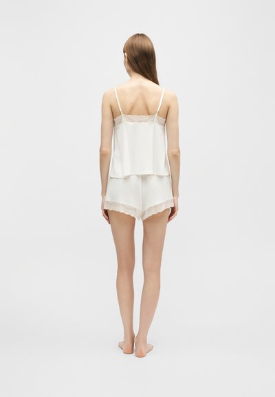 Femme debout pieds nus, dos tourné, portant un caraco blanc orné de dentelle et un short assorti sur un fond clair uni.