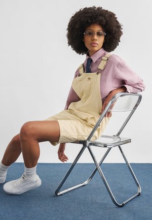 Vrouw met afro-kapsel, draagbaar zonnebril, roze shirt, beige tuinbroek, witte sneakers, zittend zijwaarts op een transparante vouwstoel op een blauw tapijt.