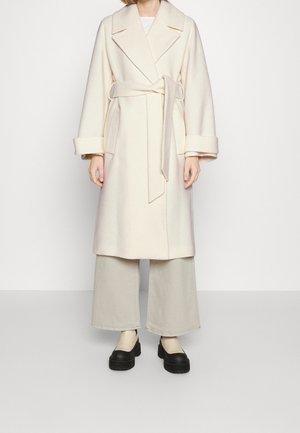 Personne portant un long manteau crème portefeuille avec de larges revers, des manches retroussées, un pantalon à jambes larges et des chaussures à plateforme noires épaisses.
