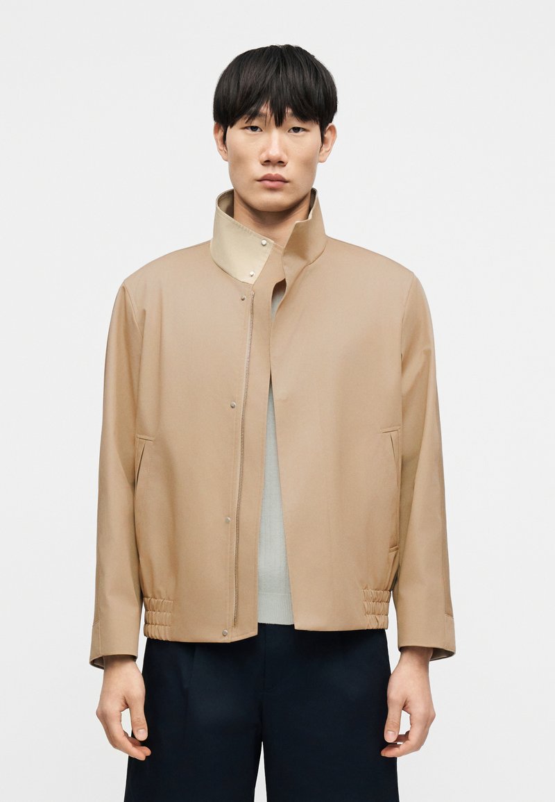 Veste légère beige avec un col montant, une fermeture à pression à l'avant et un effet froncé à l'ourlet. Texture lisse, design minimaliste.