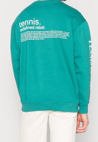 Sweatshirt teal avec texte blanc au dos, présentant des graphismes en gros caractères et des informations imprimées sur le tennis, avec des poignets et un ourlet en côtelé.