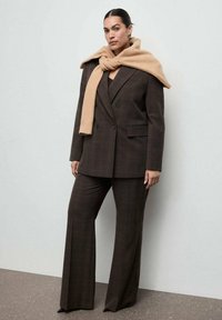 Blazer doppiopetto a quadri marroni, pantaloni a gamba larga abbinati e sciarpa beige drappeggiata sulle spalle. Tessuto liscio e design su misura.