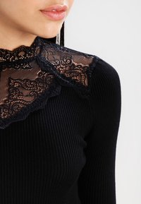 Černý žebrovaný top s vysokým krajkovým límcem zdobeným složitými květinovými vzory. Krajka je jemně propracovaná a doplňuje hladkou strukturu pod ní.