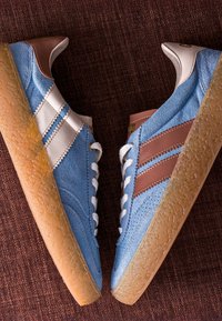Paire de baskets en daim bleu avec des rayures marron et blanches et des semelles en caoutchouc marron clair texturées sur une surface en tissu foncé.