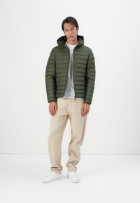 Green padded jacket with a hood, featuring horizontal quilted texture, a front zipper, and elastic cuffs; worn over a gray shirt and beige pants.